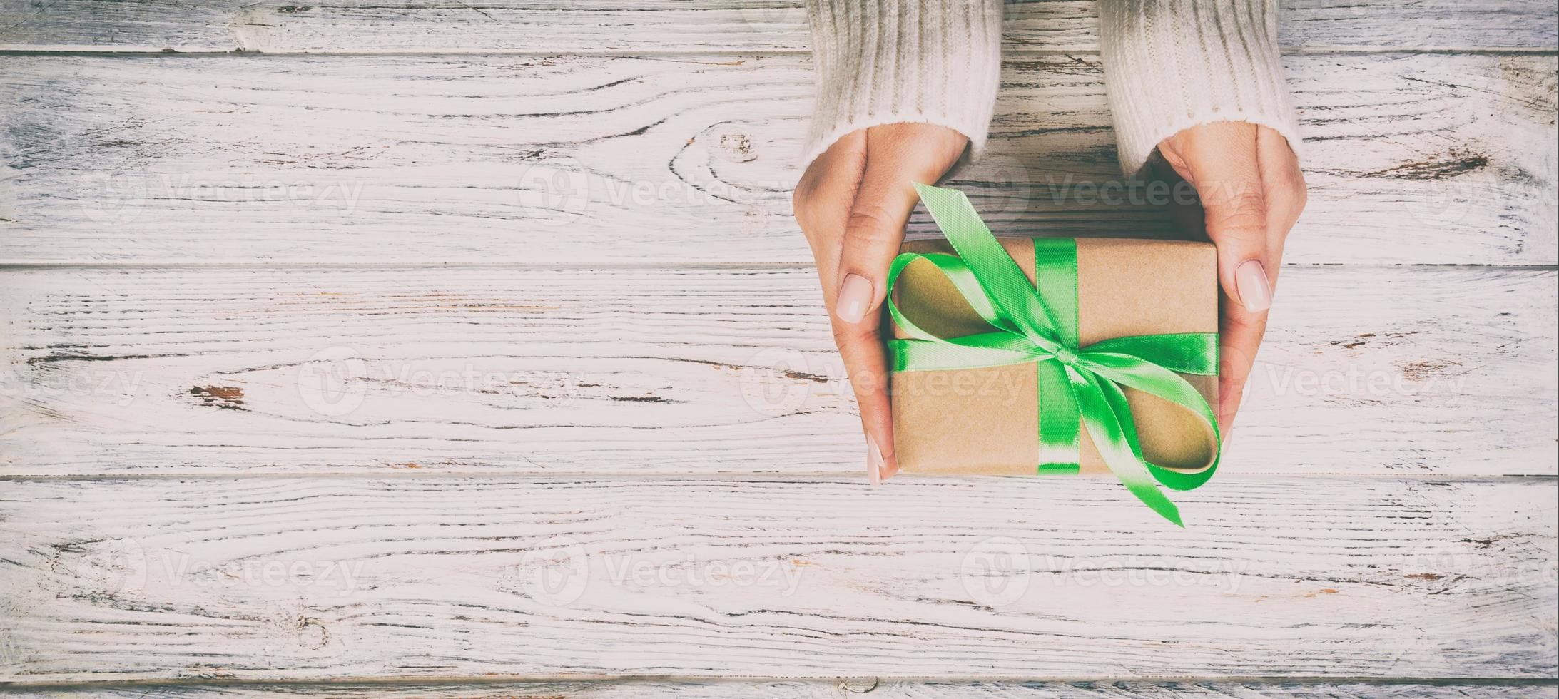 Woman hands give wrapped valentine or other holiday handmade present in paper with green ribbon. Present box, decoration of gift on wooden table, Top view banner with copy space for you design photo