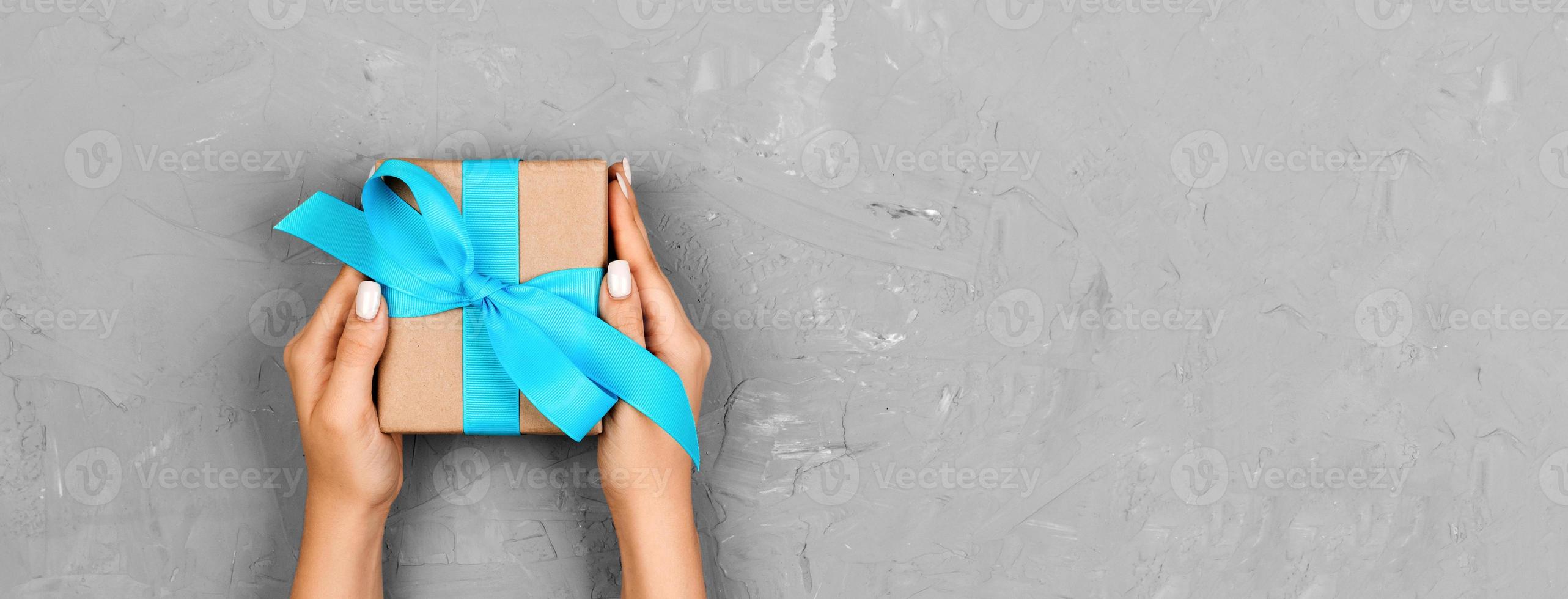 Woman hands give wrapped valentine or other holiday handmade present in paper with blue ribbon. Present box, decoration of gift on white table, top view with copy space. Banner photo