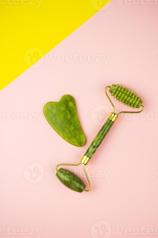 herramientas de masaje facial verde gua sha. rodillo de jade cuarzo verde sobre fondo rosa y amarillo. Cuidado antiedad, lifting y tonificante en casa. vista superior, lugar para una inscripción. foto