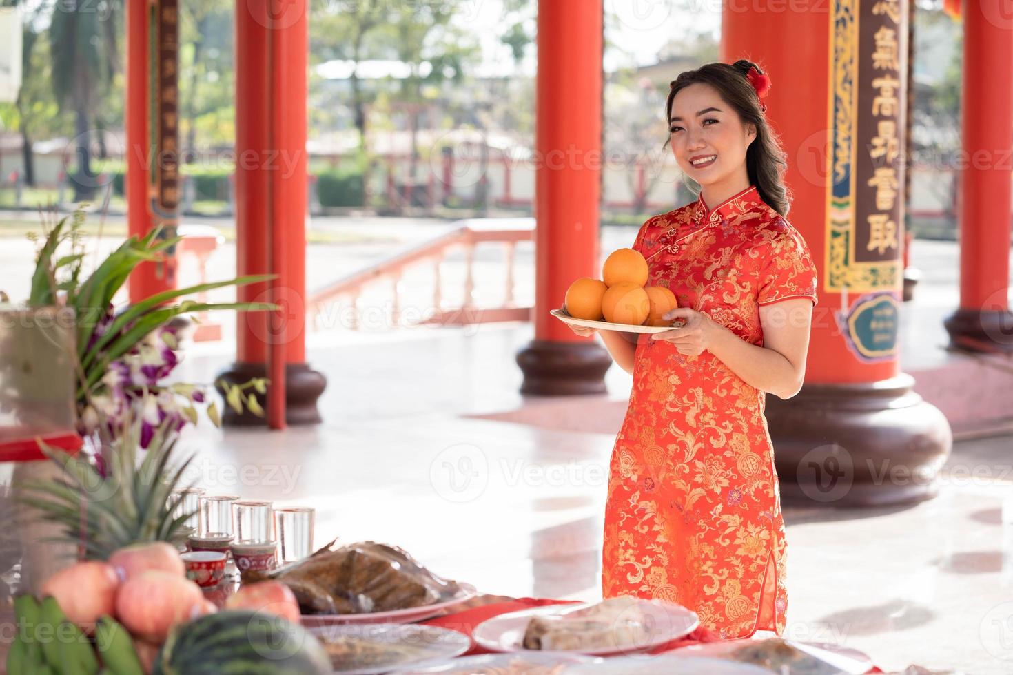 hermosa joven asiática sonriendo felizmente usando el tradicional vestido cheongsam qipao sosteniendo naranjas frescas orando por la mejor bendición y buena suerte en el templo budista chino. foto