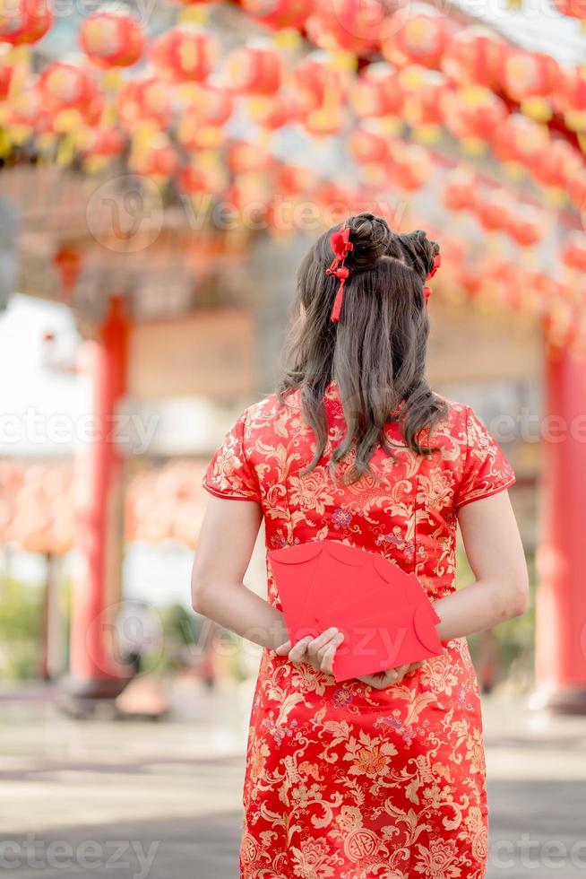 hermosa mujer asiática con vestido tradicional cheongsam qipao y parada de espaldas sosteniendo ang pao, sobres rojos en el templo budista chino. celebrar el año nuevo lunar chino. foto