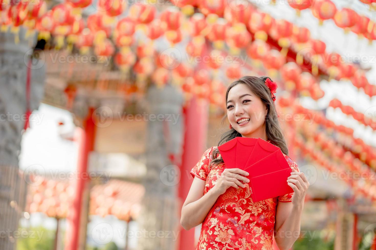 hermosa mujer asiática sonriendo felizmente sosteniendo ang pao, sobres rojos usando cheongsam luciendo confiada en el templo budista chino. celebrar el año nuevo lunar chino, vacaciones de temporada festiva foto