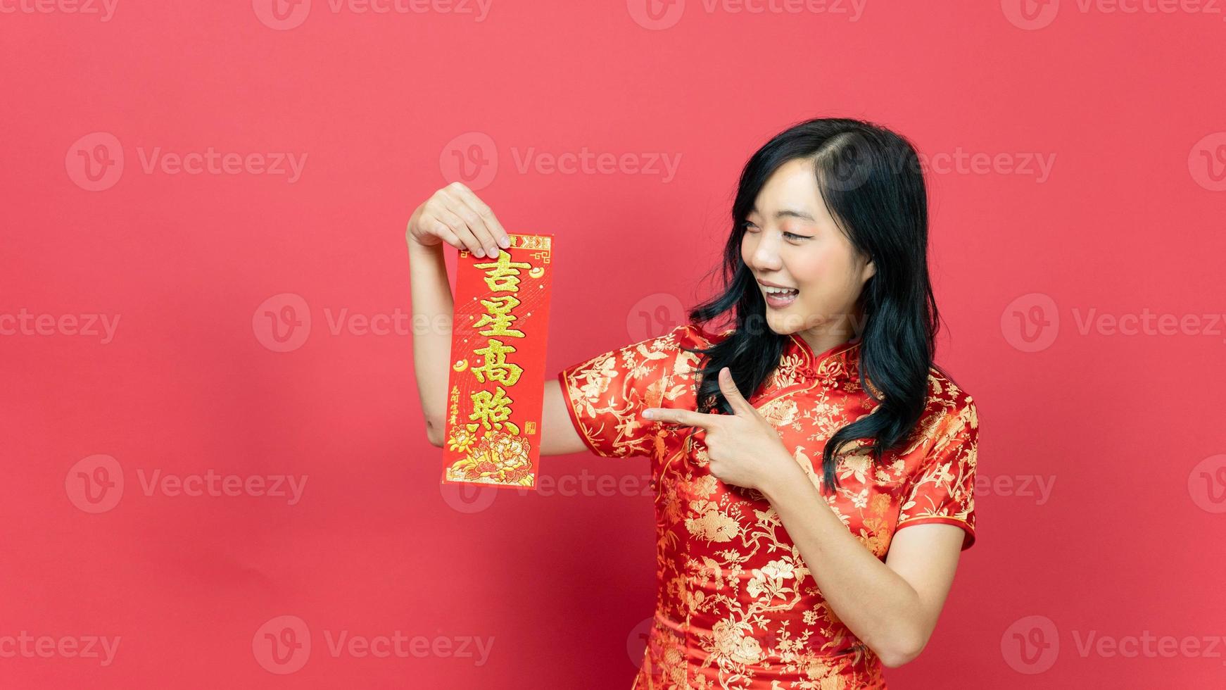 Beautiful woman wearing traditional cheongsam qipao  costume holding and pointing blessing fortune card isolated on red background. Chinese text means great luck great profit photo