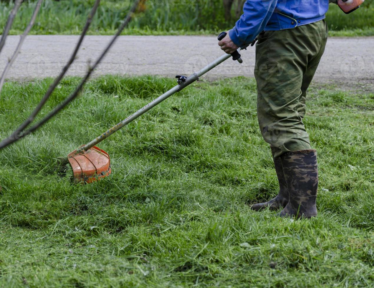 A close-up of a worker in protective clothing, gloves, rubber boots with a gas mower on the front lawn. A man mows grass with dandelions on a warm sunny spring day. photo