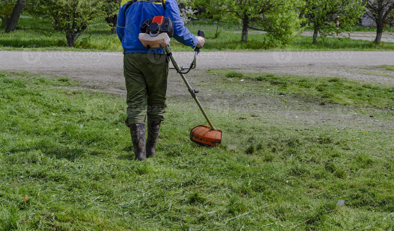 A close-up of a worker in protective clothing, gloves, rubber boots with a gas mower on the front lawn. A man mows grass with dandelions on a warm sunny spring day. photo