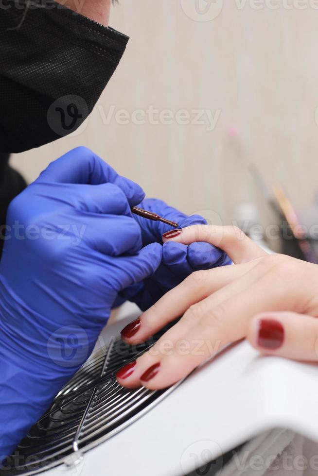 Master in protective gloves during a manicure at beauty salon. Master manicurist varnishes the marsala gel on the nails of a female client. The concept of beauty and health. photo