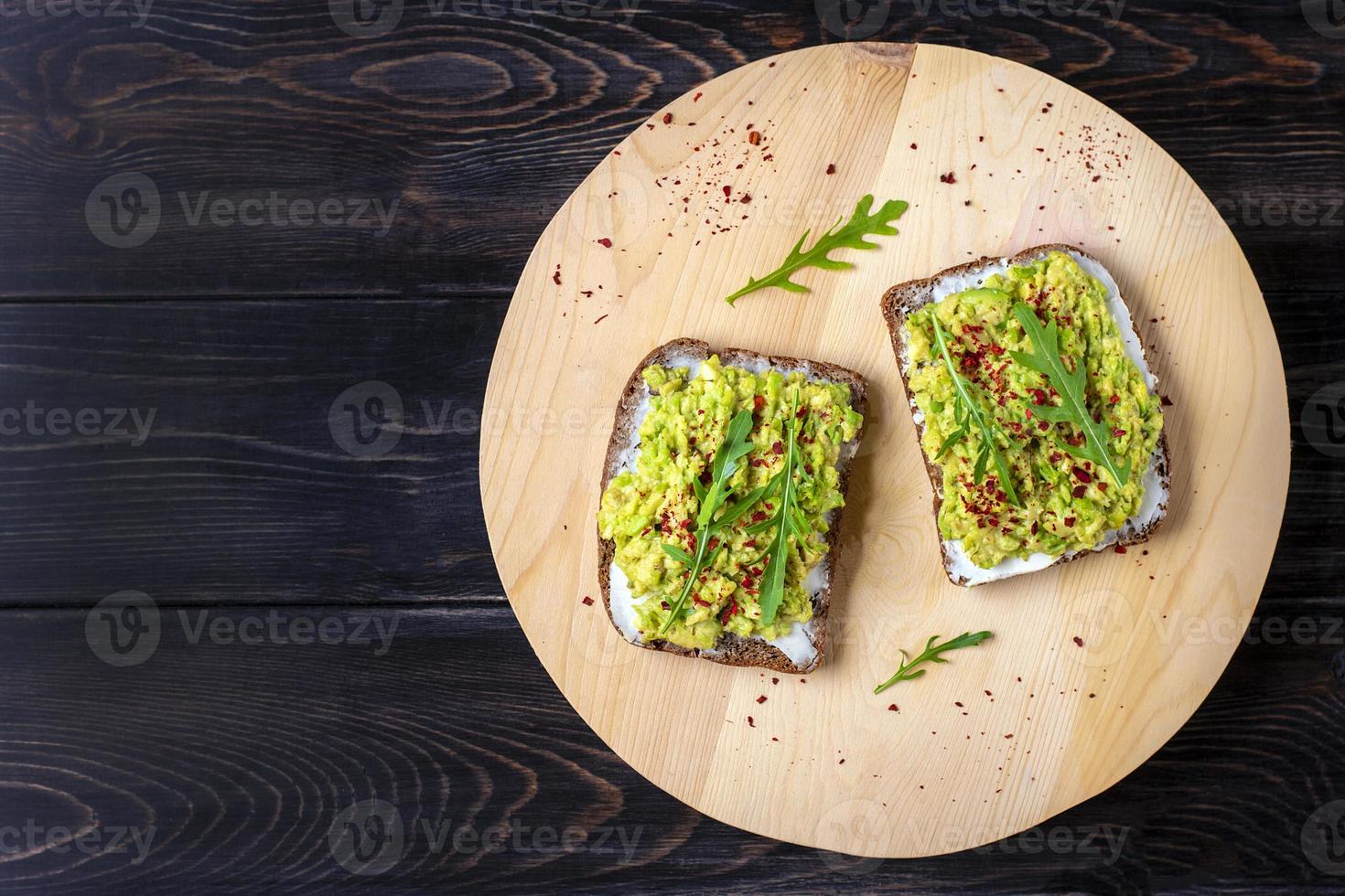 Sándwiches para el desayuno: rebanada de pan oscuro de grano entero, queso crema, guakomole, adornado con rúcula, pimentón en la mesa de madera, vista superior plana, concepto de comida saludable hecha en casa foto