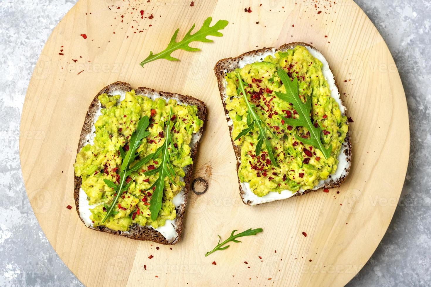 Sándwiches para el desayuno: rebanada de pan oscuro de grano entero, queso crema, guakomole, adornado con rúcula, pimentón en la mesa de madera, vista superior plana, concepto de comida saludable hecha en casa foto