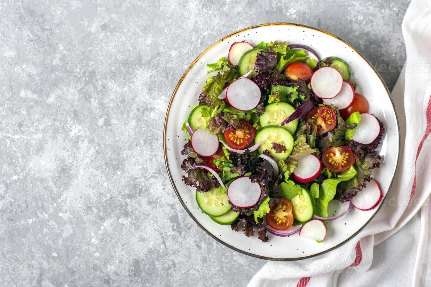 Ensalada vegetal saludable de tomates cherry, rodajas de pepino, hojas de lechuga verde y morada, cebollas y aceite de oliva en un plato sobre una mesa de hormigón vista superior dieta plana, menú mediterráneo comida vegana foto