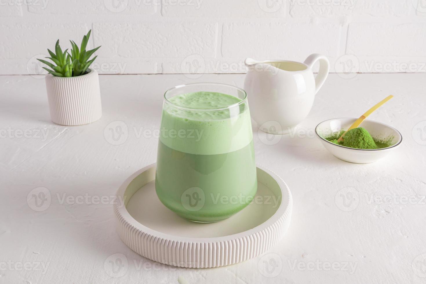 a large glass of healthy matcha latte tea with whipped milk on a round ceramic tray opposite a white brick wall. a cheerful morning without coffee. photo