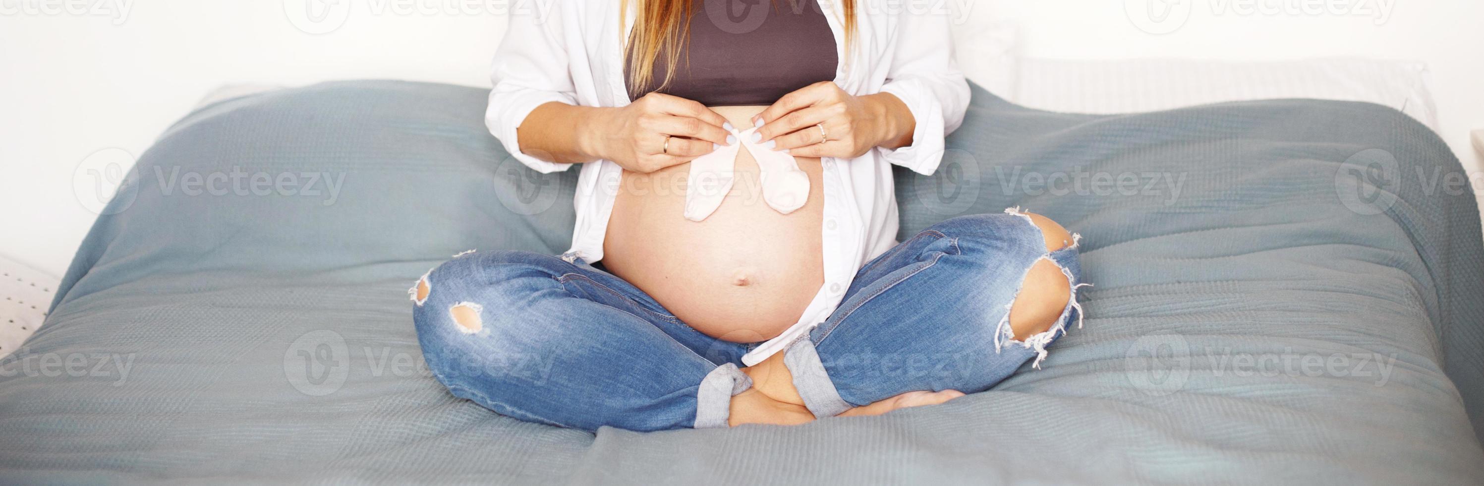 girl sitting on bed in bedroom and holding pink socks near her pregnant belly photo