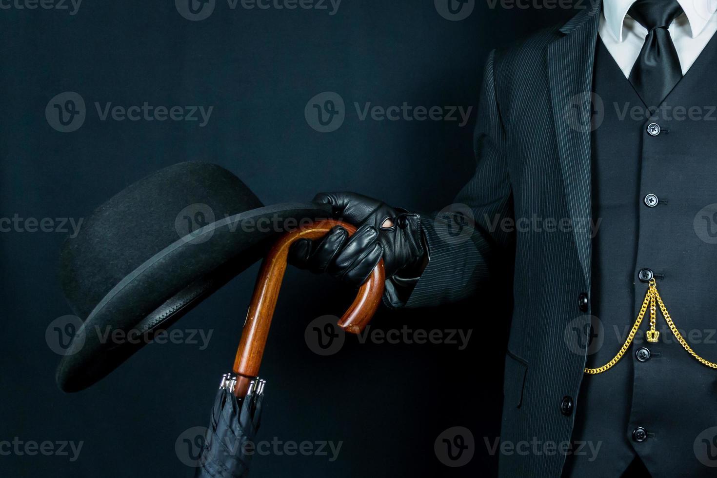 Man in Dark Suit and Leather Gloves Holding Bowler Hat and Umbrella on Black Background. Concept of Classic and Eccentric British Gentleman. photo