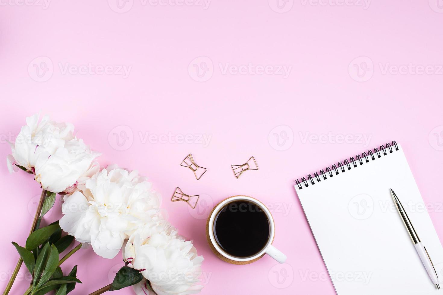 Coffee mug, empte notebook and white peony flowers on pink table background. Top view, flat lay photo