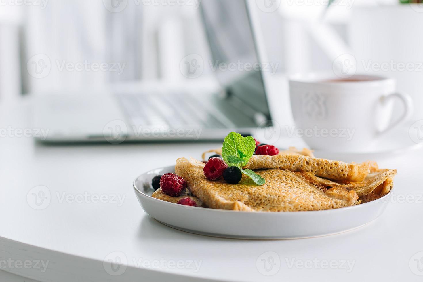 Delicious Breakfast. White desktop table with laptop and fresh crepes pancakes, cup of tea. Freelancer concept. Healthy breakfast food. photo