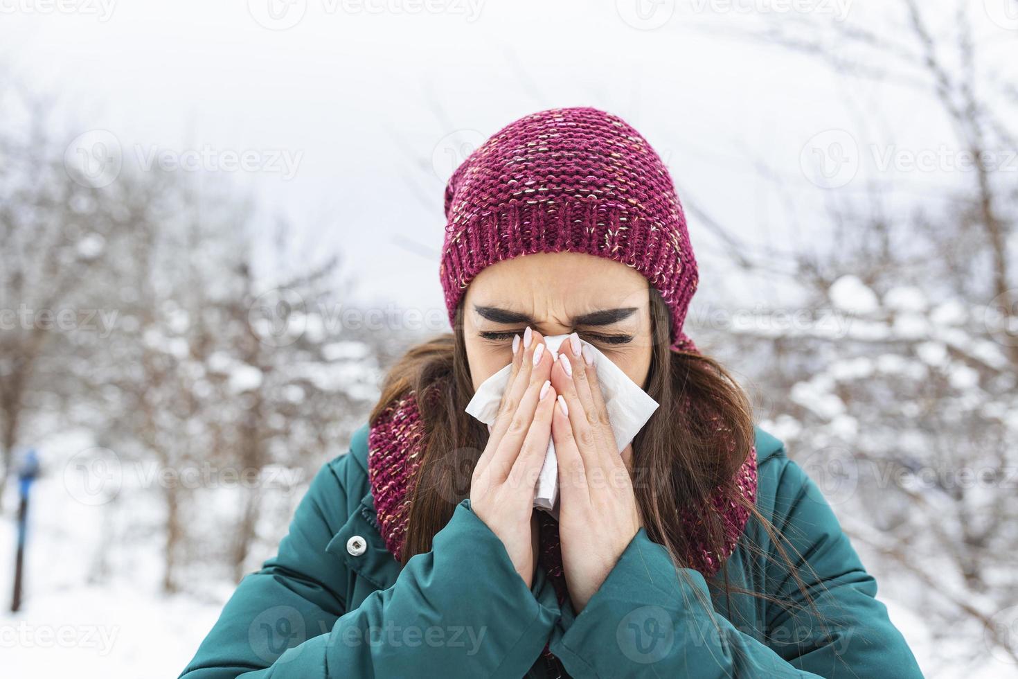 Young woman sick and sneeze into tissue paper. Girl blowing nose outdoors.Catching cold in winter. Upset gloomy woman with ill expression sneezes and has running nose photo