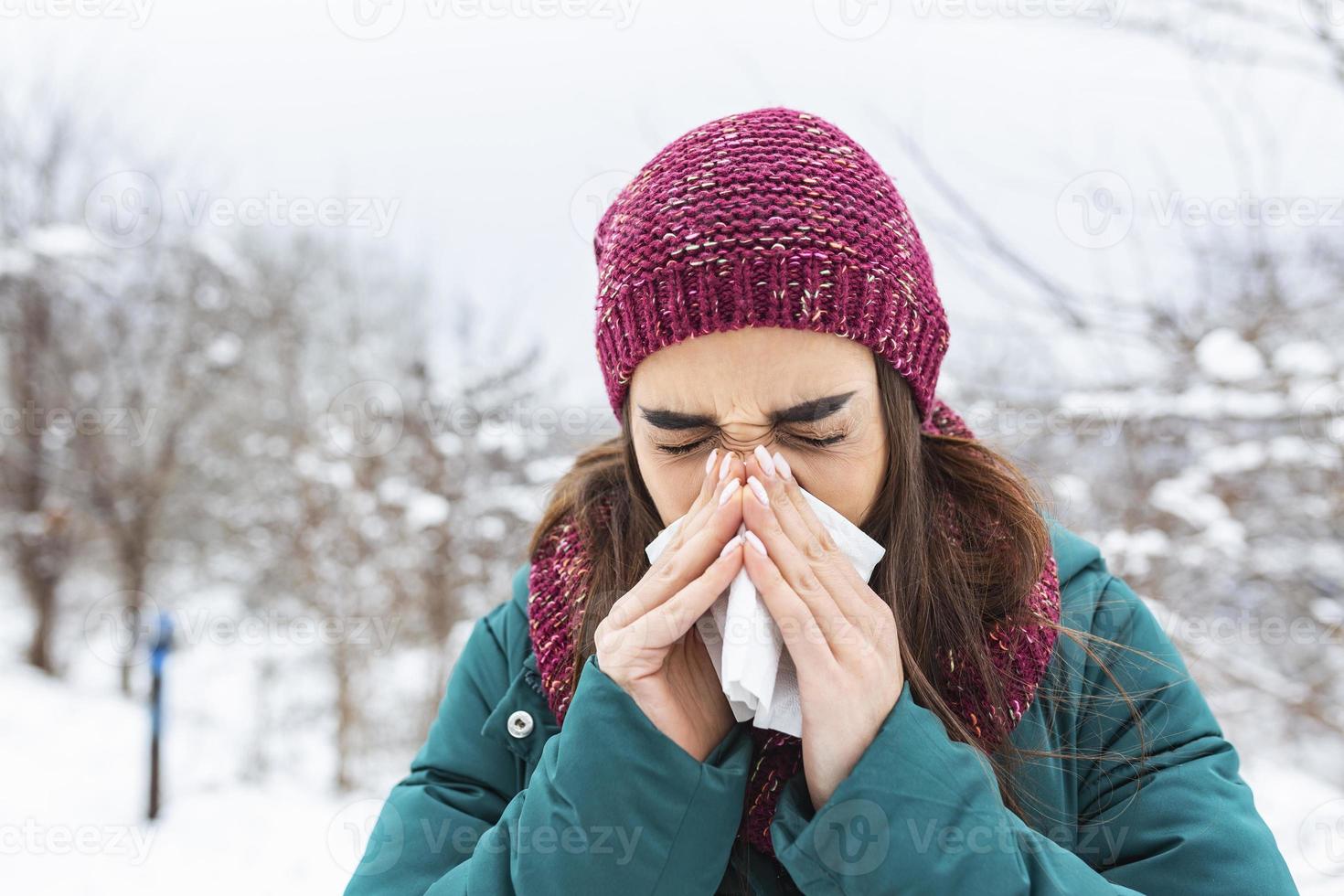 Young woman sick and sneeze into tissue paper. Girl blowing nose outdoors.Catching cold in winter. Upset gloomy woman with ill expression sneezes and has running nose photo