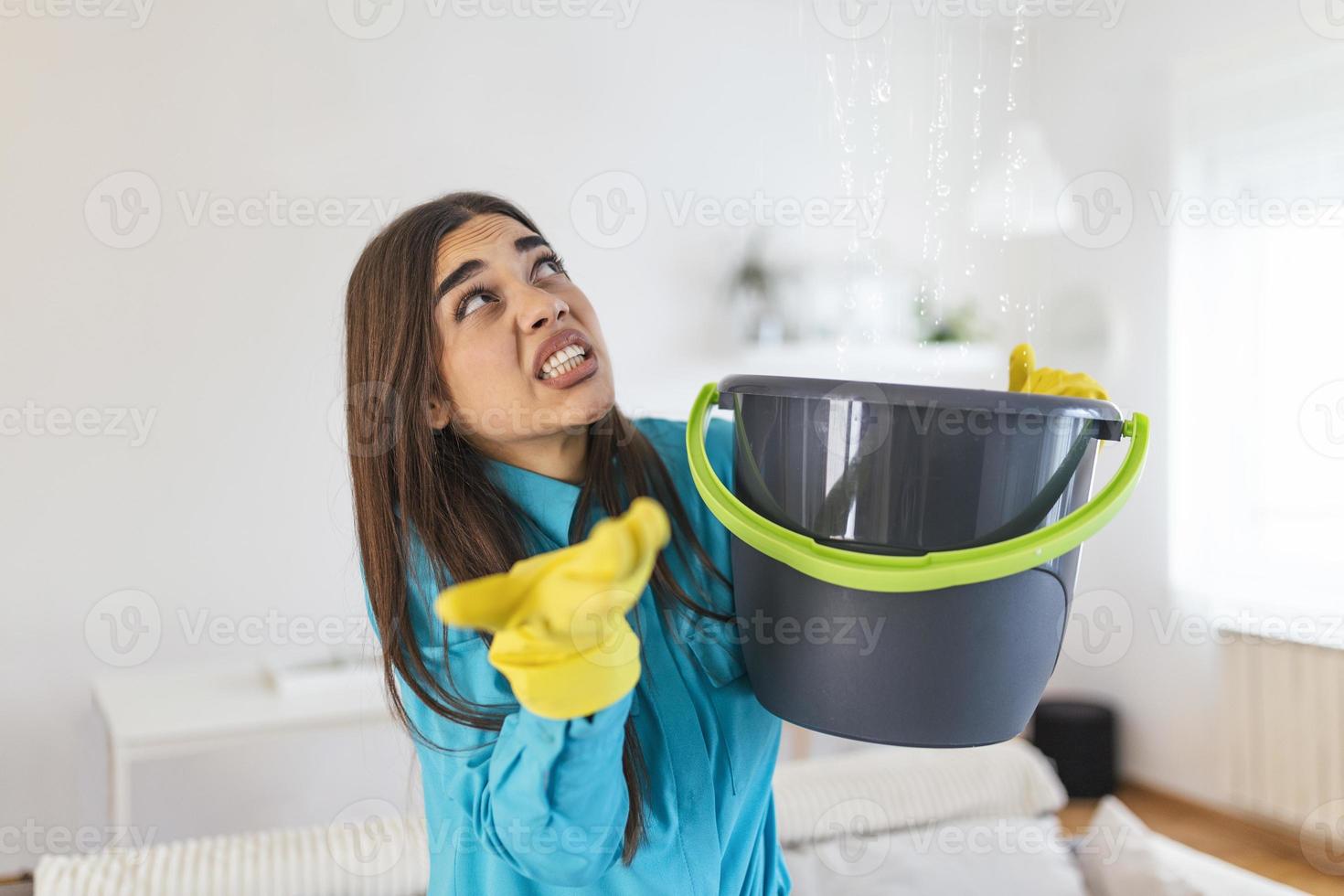 Shocked Woman Looks at the Ceiling While Collecting Water Which Leaks in the Living Room at Home. Worried Woman Holding Bucket While Water Droplets Leak From Ceiling in Living Room photo