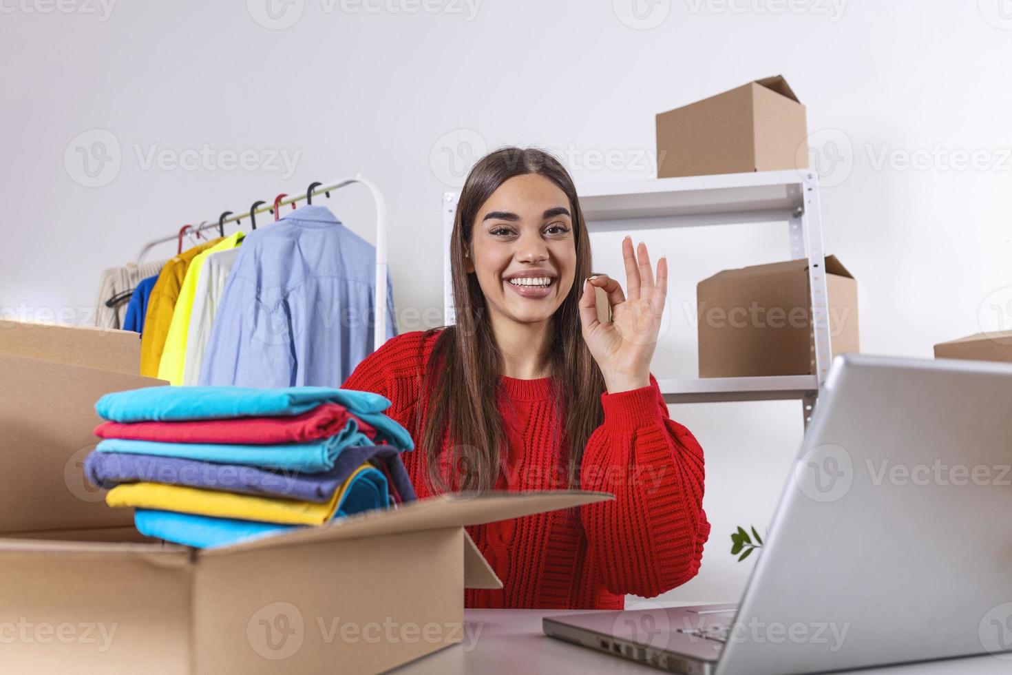 Woman packing item that she sells online. Working woman at online shop. She wearing casual clothing and checking on laptop address of customer and package information. Beautiful girl showing OK photo