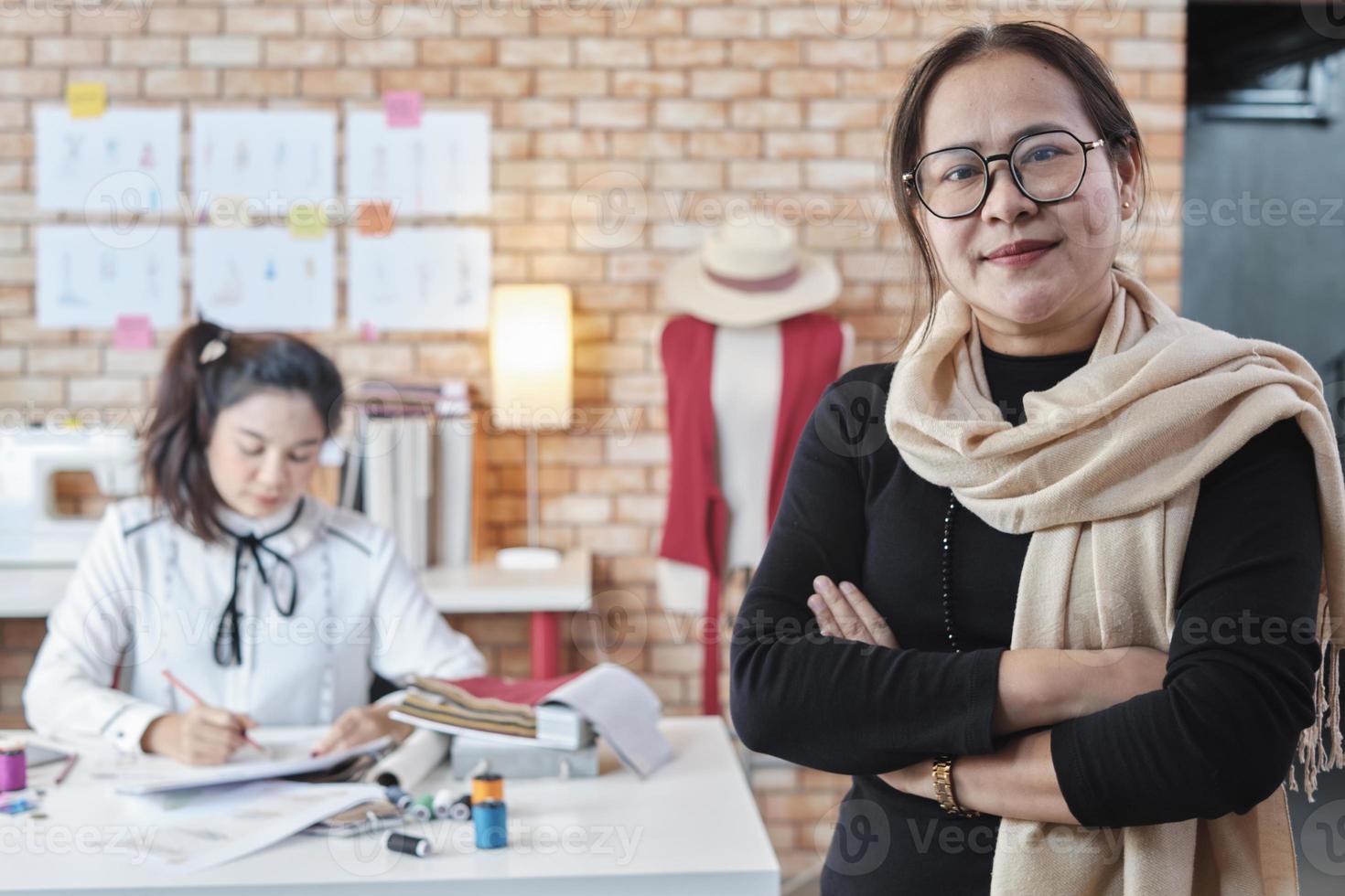 Asian mature female fashion designer looks at camera and smiles in studio, arms crossed, working with creative and sewing for dress design collection, professional boutique tailor SME entrepreneur. photo