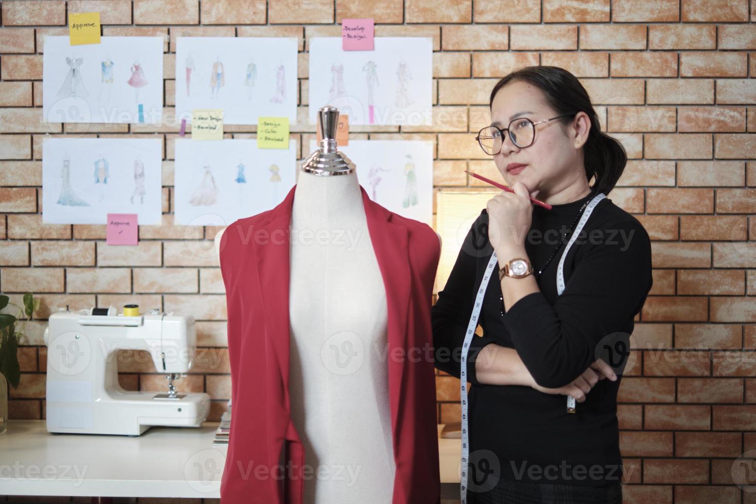 Asian mature female fashion designer stands near dressing puppet, thinking and contemplating for design ideas in studio, works with clothing collection, professional boutique tailor SME entrepreneur. photo