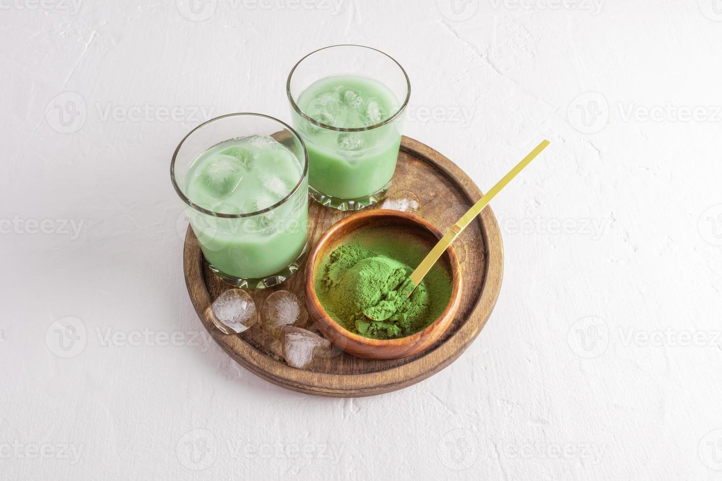 two glass of matcha latte tea with ice cubes on a wooden tray with a bowl of green tea powder . a health-improving invigorating agent. photo