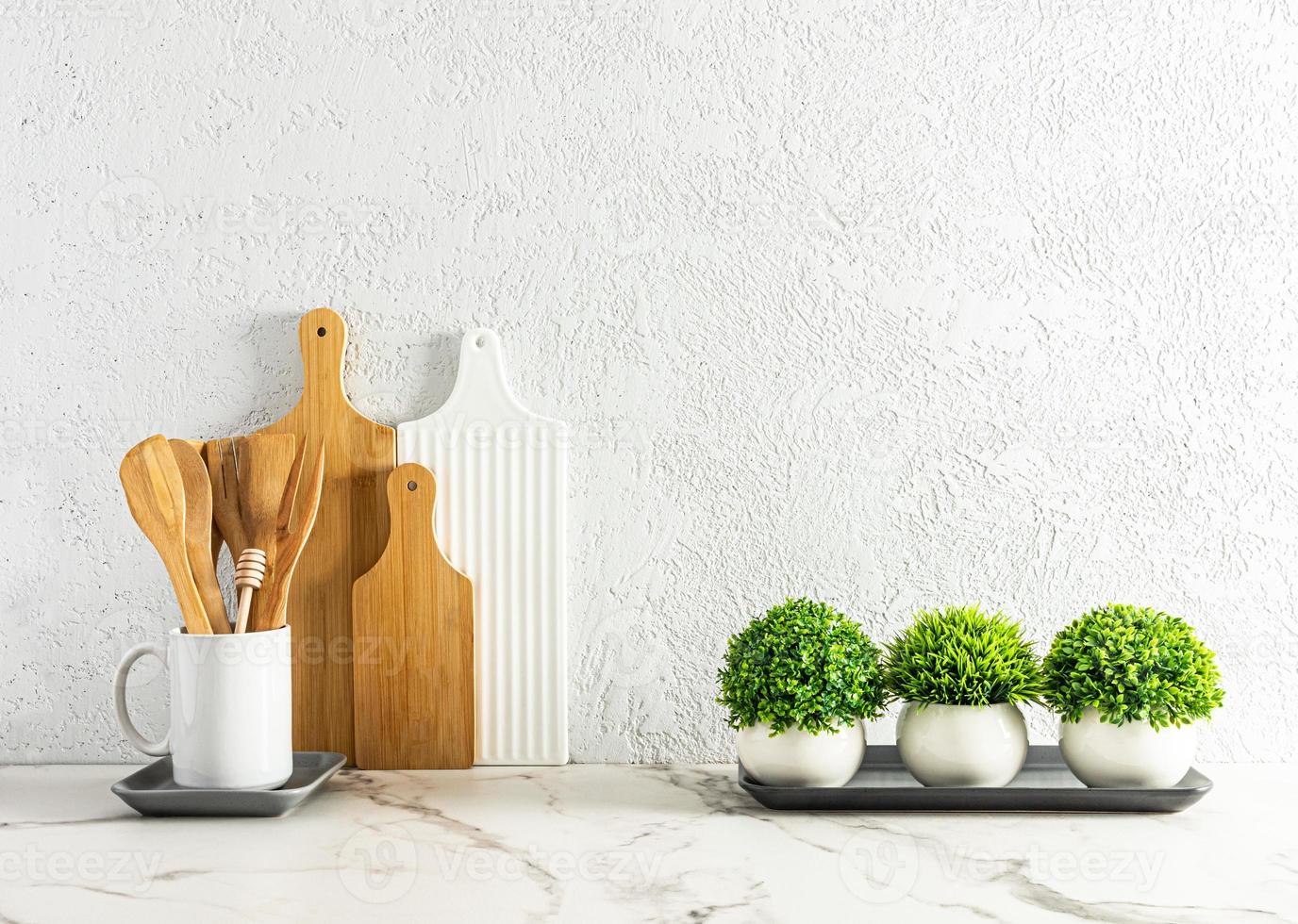 elegante fondo blanco de cocina con varias tablas de cortar y cucharas de cocina, plantas verdes en macetas sobre una encimera de mármol. foto