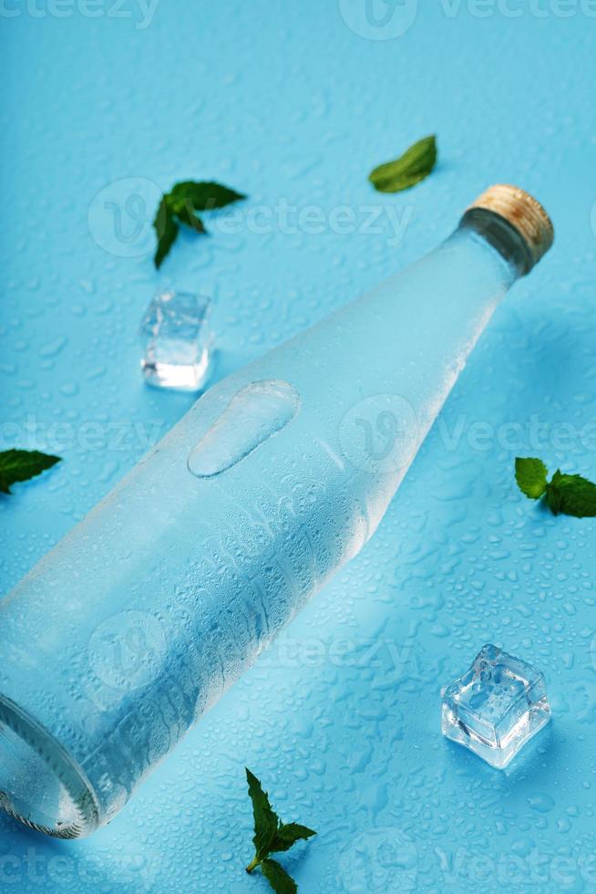 una botella de agua helada, cubitos de hielo, gotas y hojas de menta sobre un fondo azul. foto