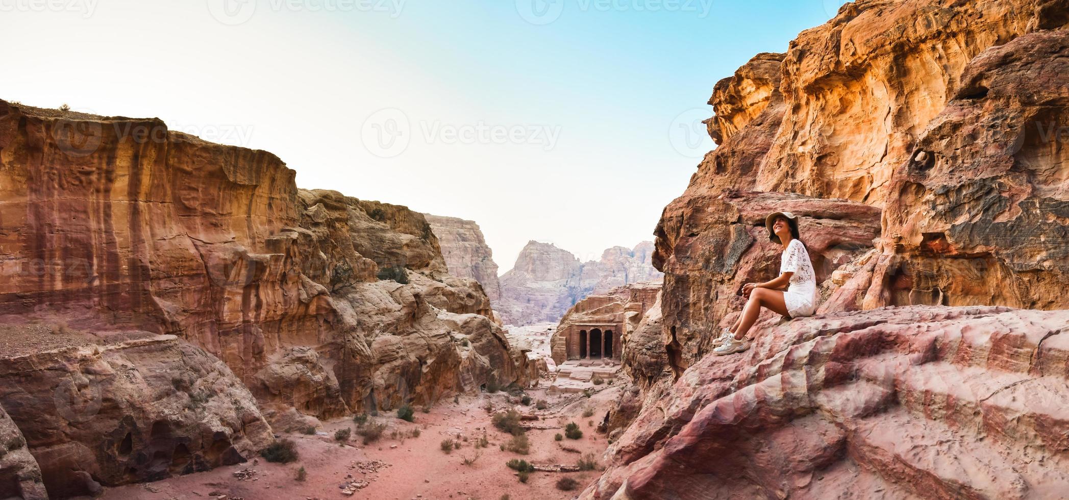 Caucasian young female tourist sit and smile while looking to beautiful Petra ancient city carvings. Famous visit Jordan attraction destination. Petra archeological site panorama photo