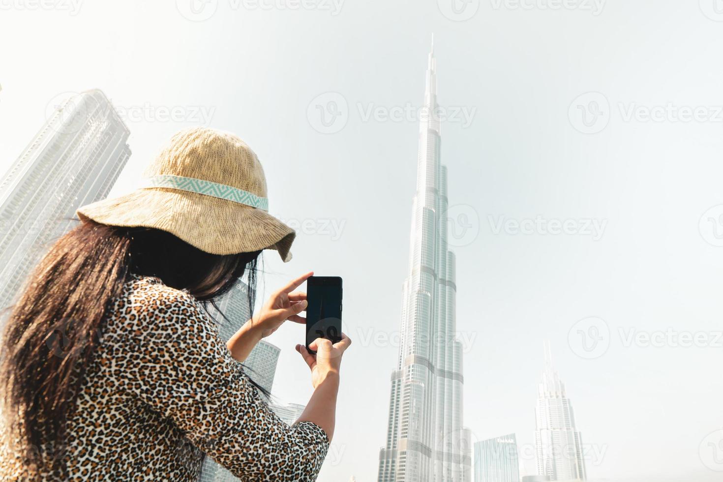 Young caucasian tourist woman takes mobile photo of Burj Khalifa with smartphone in sunny hazy sky day