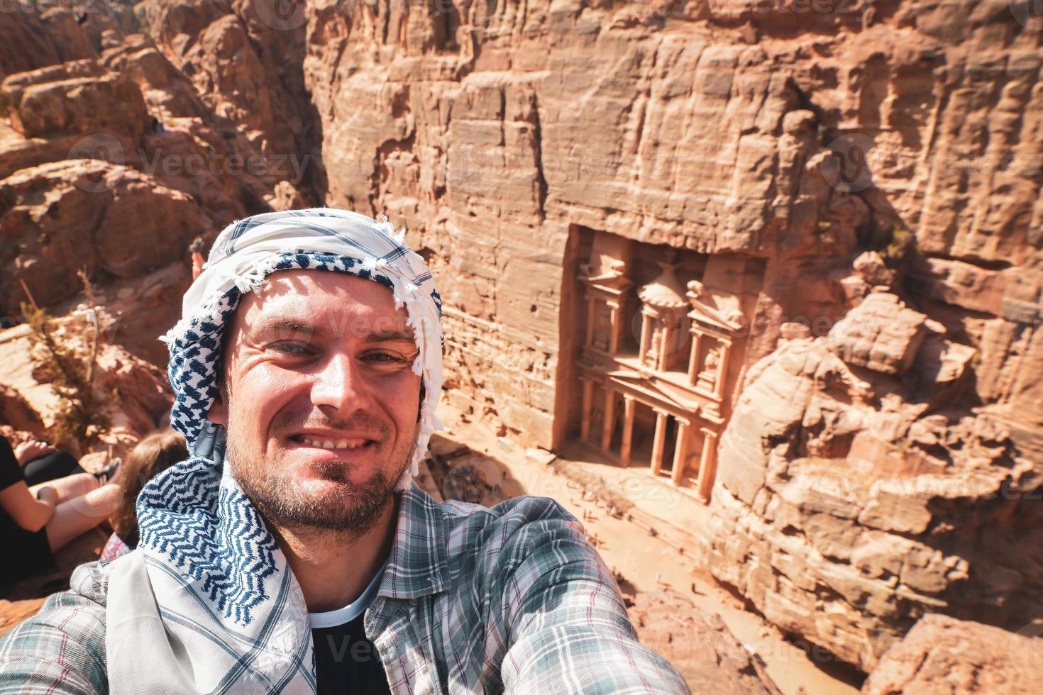 Caucasian male tourist stand on viewpoint in Petra ancient city over Treasury or Al-khazneh take smartphone photo smile. Jordan, one of seven wonders. UNESCO World Heritage site.