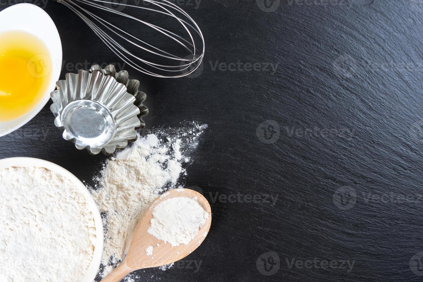 Baking ingredients flour, eggs, milk, bakeware on black slate background with copy space photo