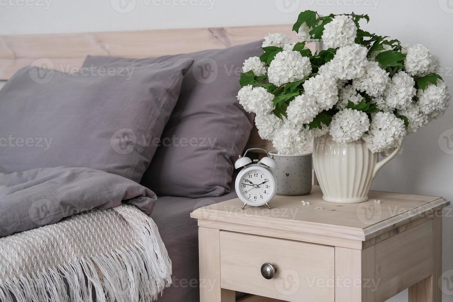 A cup of coffee, an alarm clock and a vase of white flowers on the bedside table. Tea time, coffee time photo