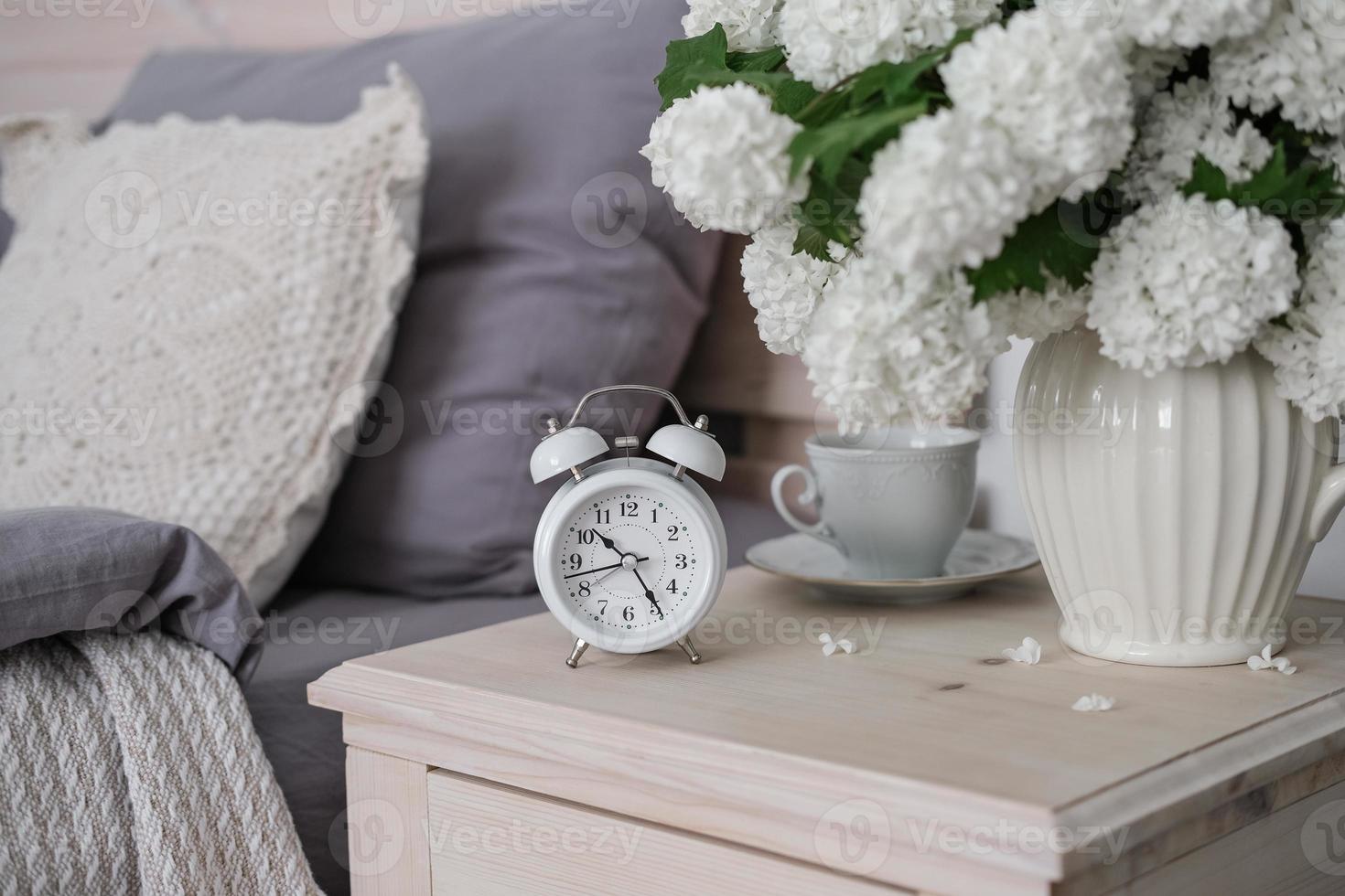 An alarm clock, a cup of tea and a vase of white flowers on the bedside table. Tea time, coffee time photo