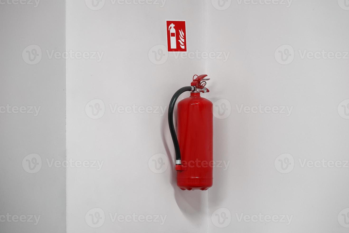 Red fire extinguisher hanged on a white wall near corner with a sticker photo