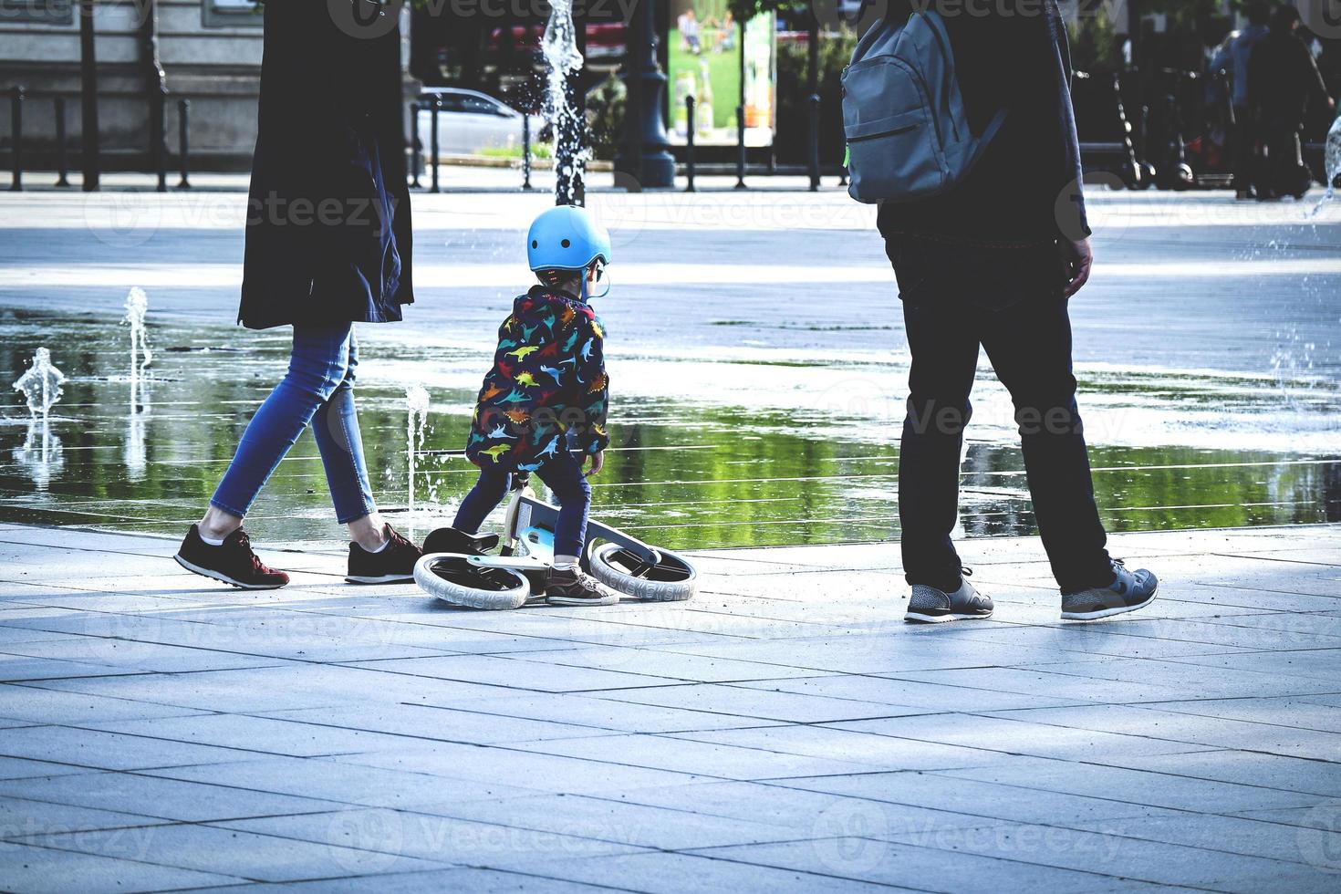 Parents with daughter walking on city square with fountain while kid in helmet is holding a scooter near huge water puddle with green trees reflection, nice family moment photo