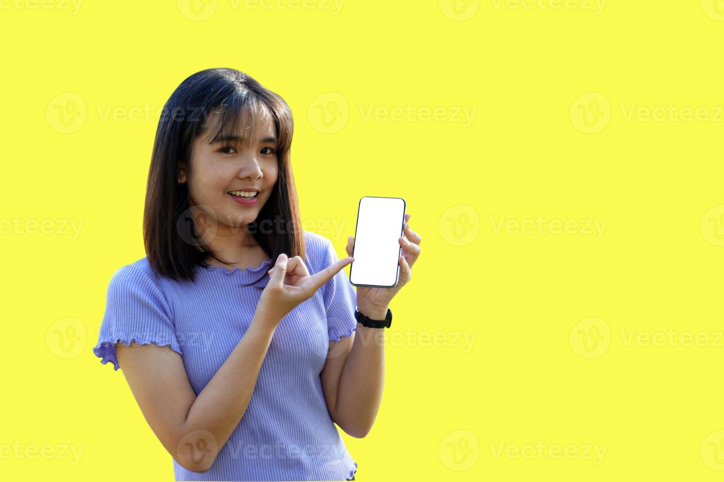 Smiling Asian woman playing and update new applications. Her hand pointed at the phone with blank screen workspace area chatting on yellow background. Soft and selective focus. photo