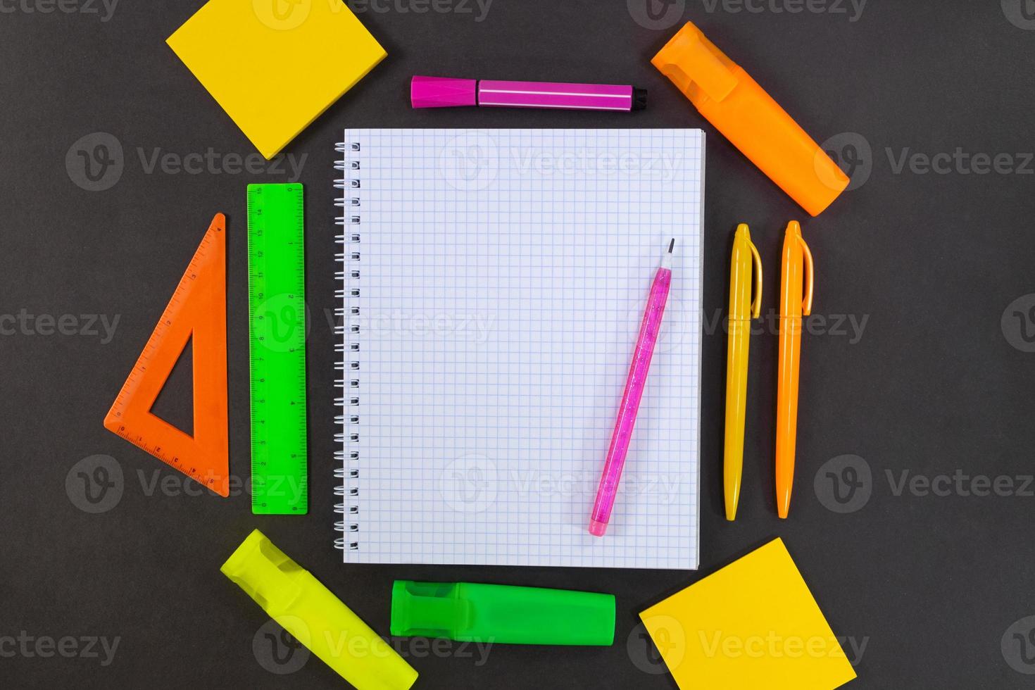 De vuelta a la escuela. en la mesa negra se encuentra el cuaderno a cuadros con lápiz para texto con espacio vacío. alrededor hay papelería verde, amarilla, rosa, naranja. reglas, rotuladores, rotuladores, bolígrafos, notas adhesivas foto