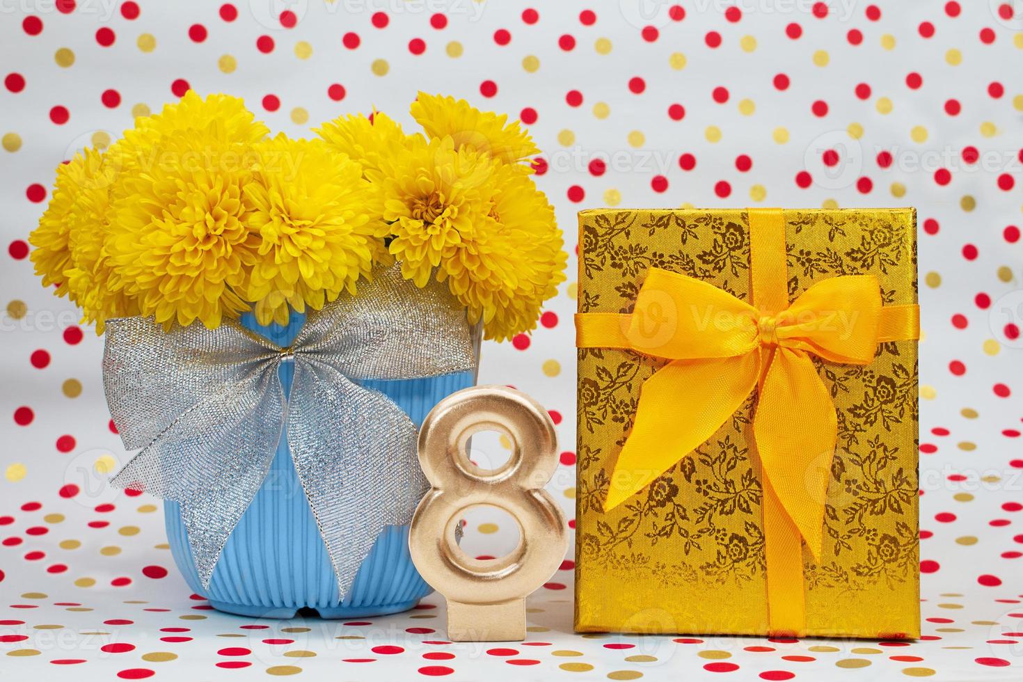 Bouquet of yellow chrysanthemums in blue pot with bow and golden gift box, number 8, behind white background with polka dots. International Women's Day, March 8, birthday photo