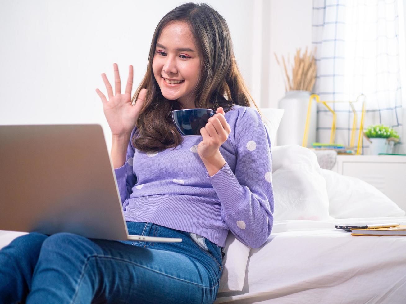 conexión de tecnología y forma moderna de pensar, la chica agitó su mano para saludar a sus amigos a través de videollamadas por Internet, se sienta y bebe café de manera relajada. concepto de distancia social foto