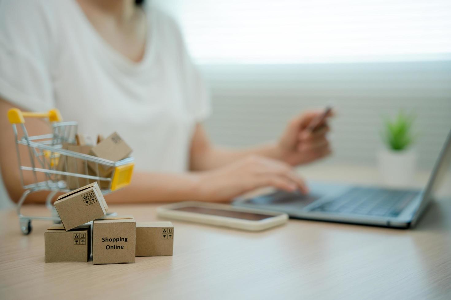 Online shopping-boxes or parcels are placed on the table and shopping carts. Blurred background, woman use credit card to make online purchases on laptop. Online service. New normal shopping delivery. photo