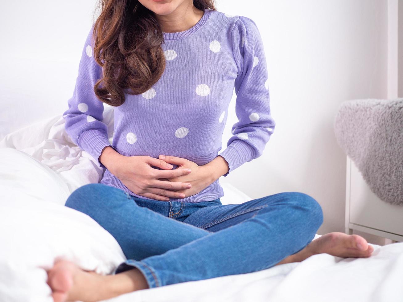 mujer sentada en la cama, usando ambas manos para agarrar el estómago, sufriendo dolor abdominal, sintiéndose torturada. concepto de menstruación foto