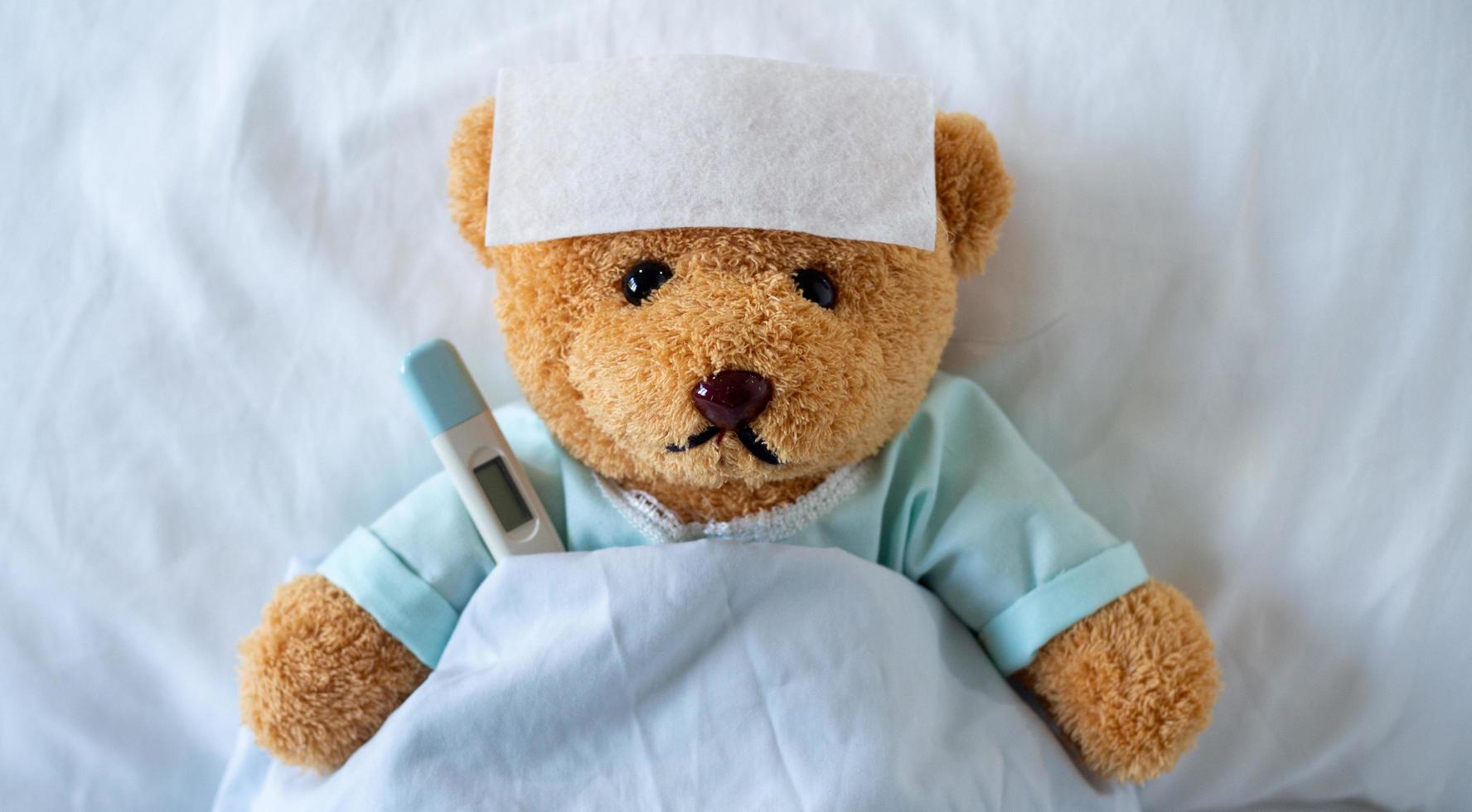The teddy bear is sick on the bed with a high fever. There is a fever reducing sheet on the forehead. photo