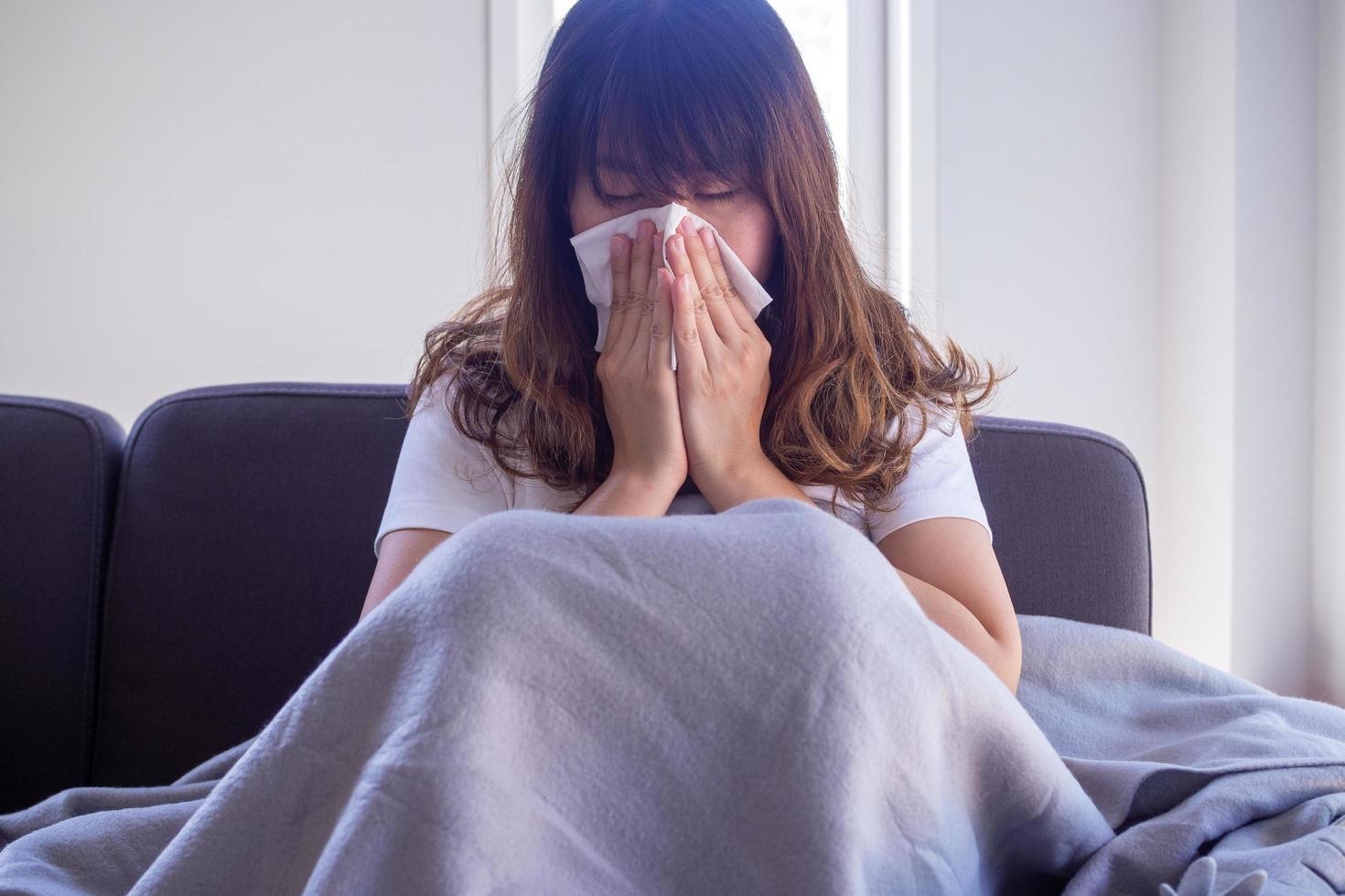 mujer de pelo largo sentada en el sofá sufre de gripe, tos y estornudos. sentarse en una cobija debido a la fiebre alta y taparse la nariz con un pañuelo de papel porque estornuda todo el tiempo. foto