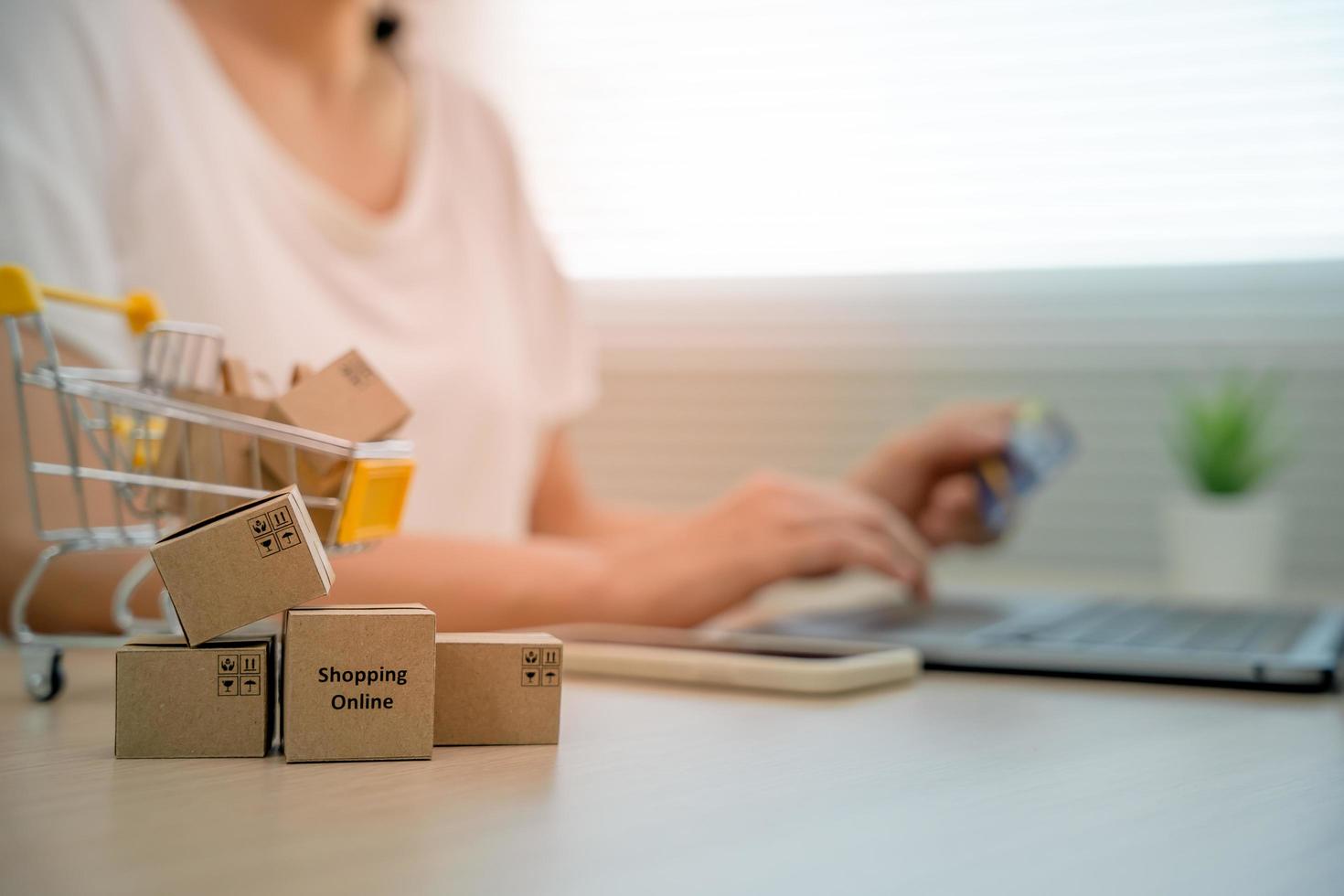 Online shopping-boxes or parcels are placed on the table and shopping carts. Blurred background, woman use credit card to make online purchases on laptop. Online service. New normal shopping delivery. photo