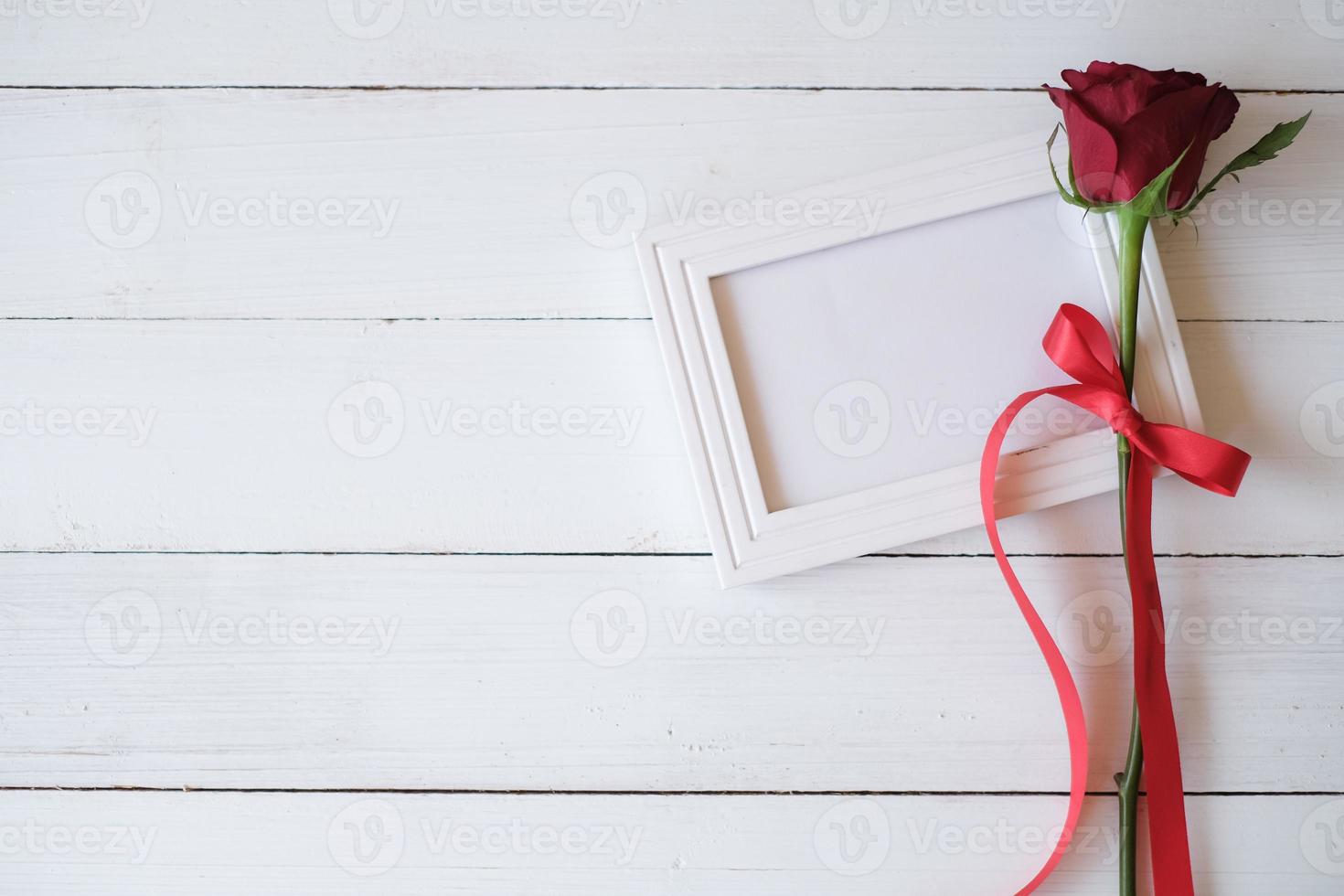 Red rose with red ribbon on a white blank photo frame isolated on white wooden background. Valentine's day, wedding, birthday and special occasion concept. Flat lay, top view. Copy space for text.
