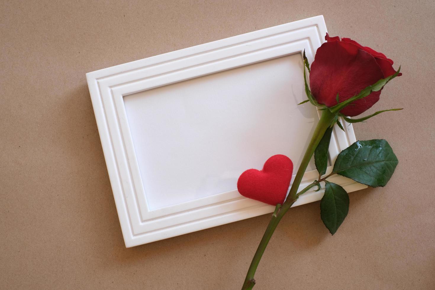 Top view of red rose and red heart laying on a white blank photo frame isolated on brown background. Valentine's day, wedding, birthday and special occasion concept. Flat lay, top view.