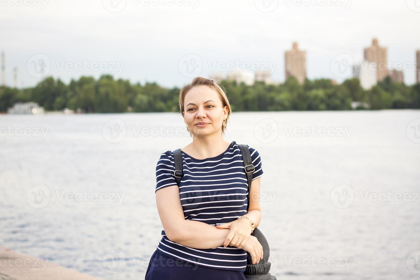 A girl in a striped jacket is standing on the river embankment, leaning on the parapet, looking into the distance. Evening walk around the city. photo