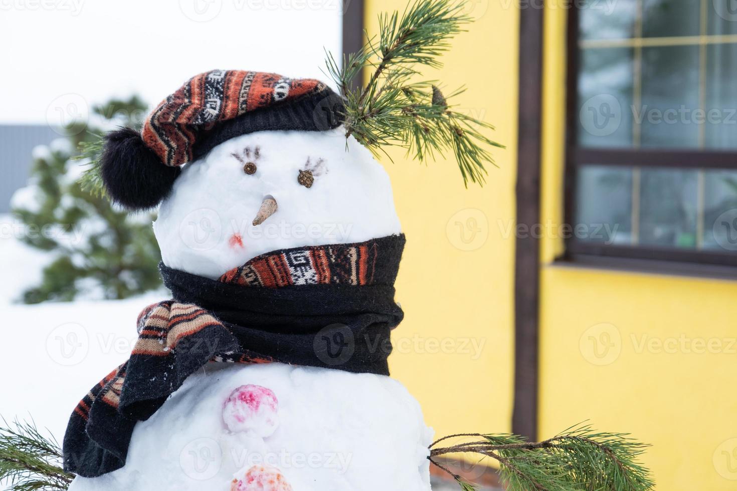 divertido muñeco de nieve con sombrero y bufanda en el fondo de una casa amarilla en el patio. invierno, entretenimiento de invierno, nevadas foto