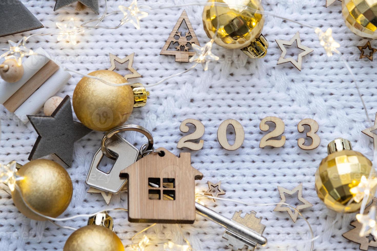 House key with keychain cottage on festive knitted background with stars, lights of garlands. Happy New Year 2023-wooden letters, greeting card. Purchase, construction, relocation, mortgage photo