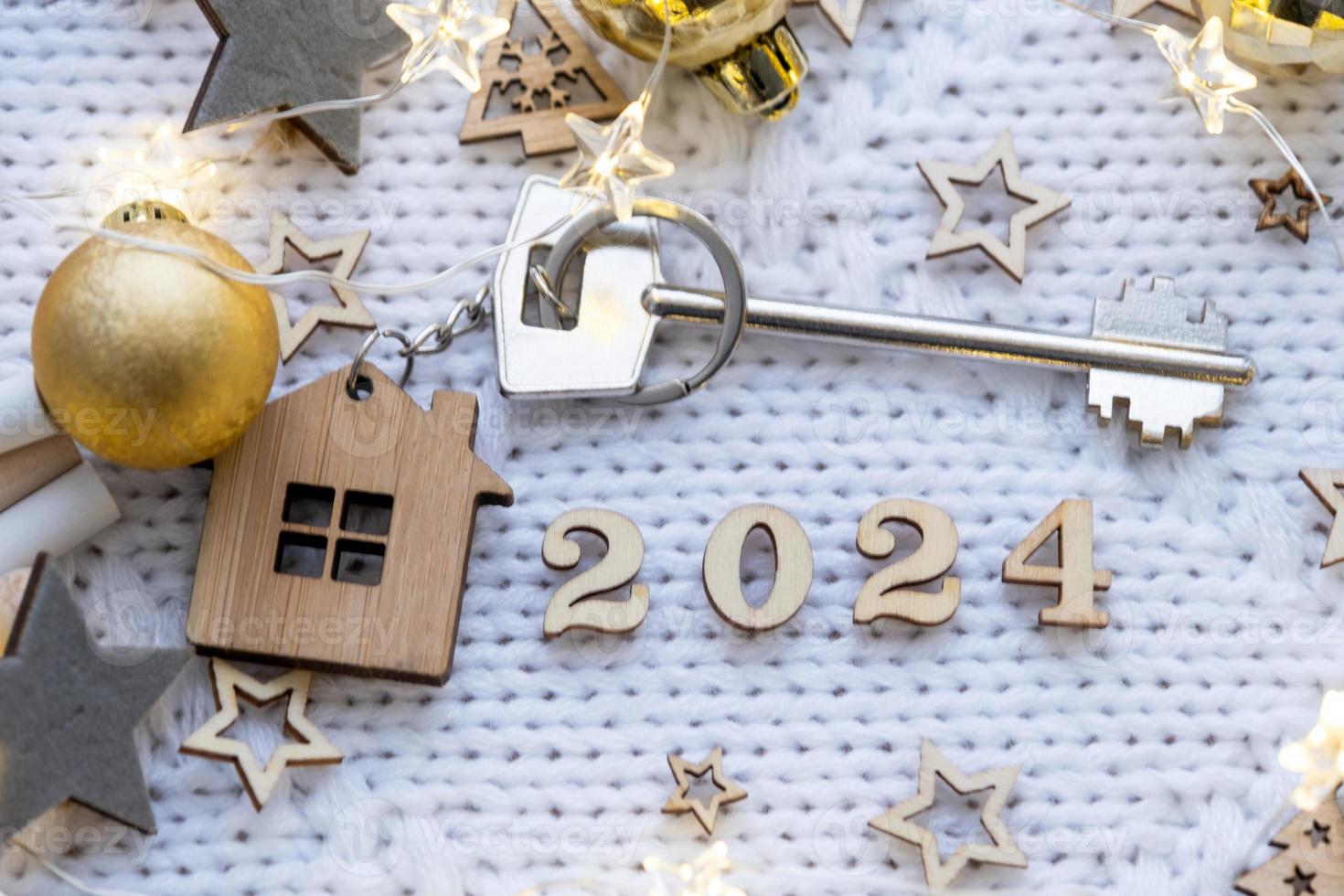 House key with keychain cottage on festive knitted background with stars, lights of garlands. Happy New Year 2024-wooden letters, greeting card. Purchase, construction, relocation, mortgage photo